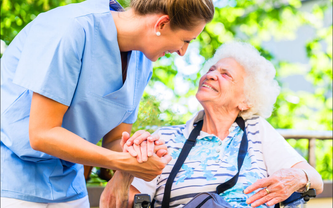 Pflegekraft hält die Hand einer pflegebedürftigen, älteren Dame im Rollstuhl. Hier wird der Beruf einer Pflegekraft deutlich.