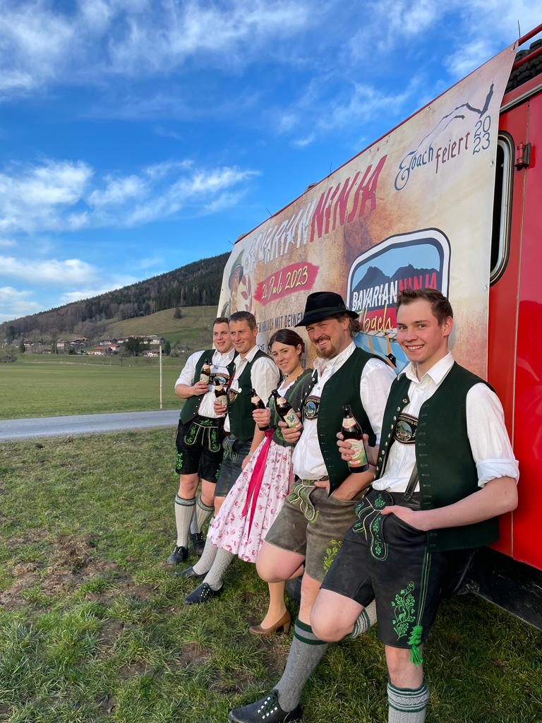 Elbach feiert den Brass Freida und Bavarian Ninja im Festzelt Elbach. Gruppe von jungen Trachtler aus Elbach stehen vor altem Feuerwehrauto mit Werbung von dem Event Bavarian Ninja.