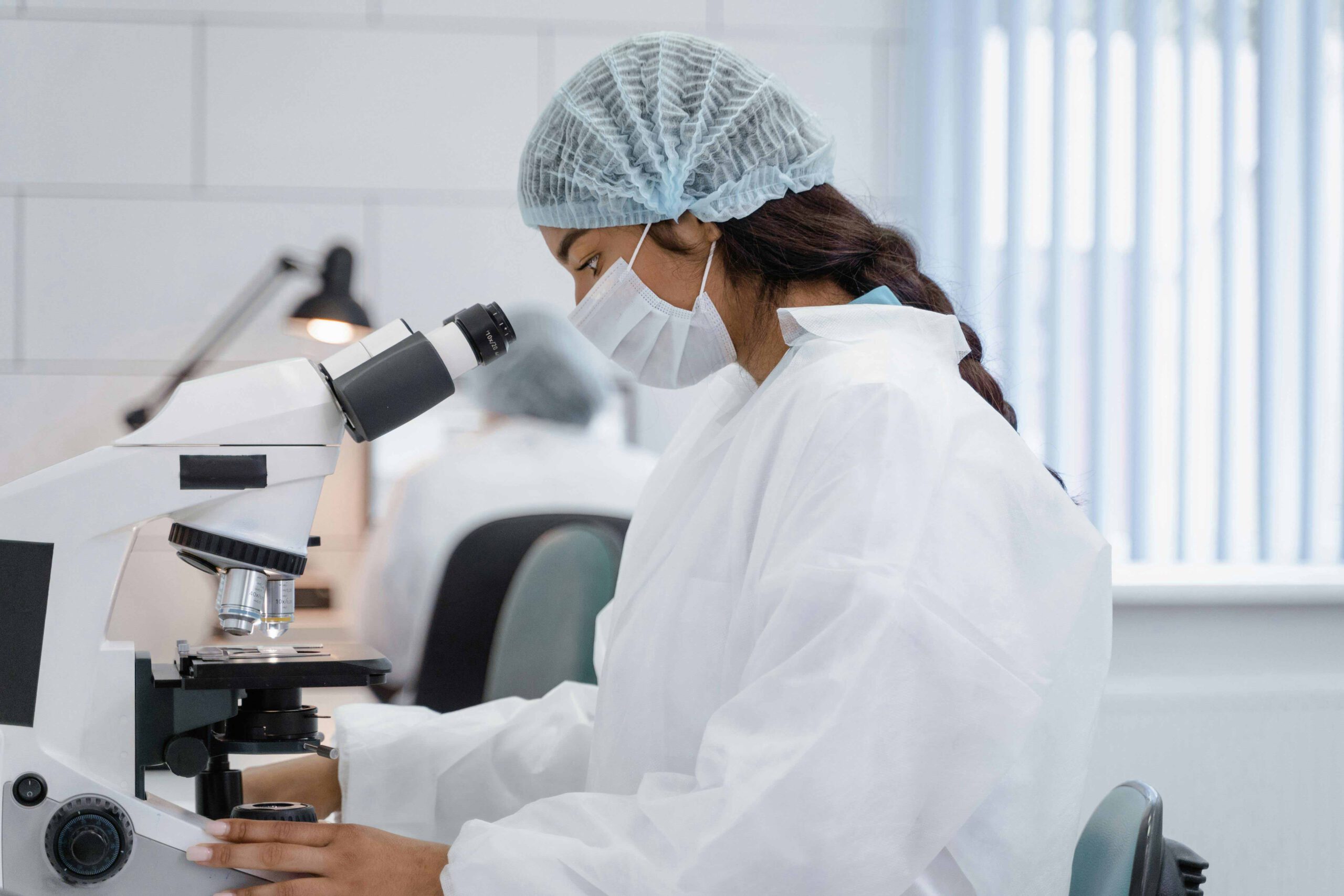Eine Frau in einem weißen Kittel und einem Haarnetz sitzt vor einem Mikroskop und untersucht eine Probe.